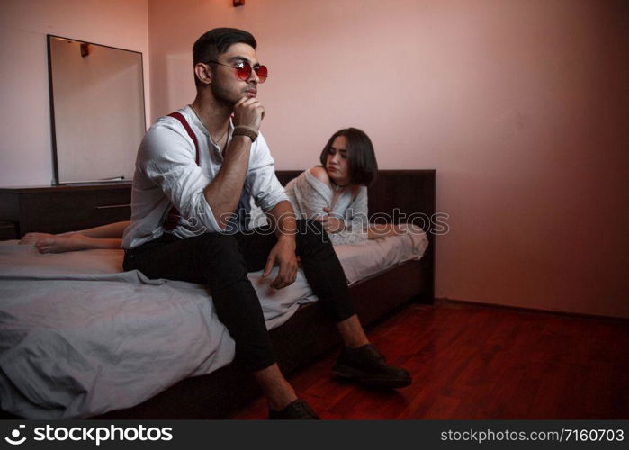 a young stylish guy in glasses sits on the edge of the bed with a thoughtful sad face. behind him lies a young brunette girl.