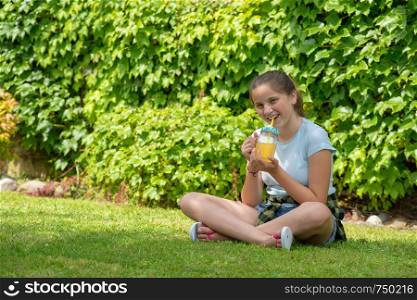 a young teenager girl sitting in the grass and drinking orange juice