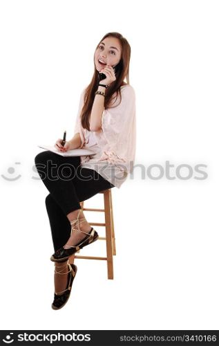 A young teenager sitting on a chair, in black tights and with long brunettehair and a notebook in her hand, on the cell phone, for white background.