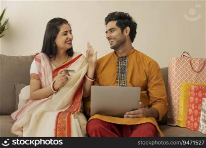 A YOUNG WOMAN HAPPILY GESTURING TO HUSBAND WHILE DOING ONLINE SHOPPING