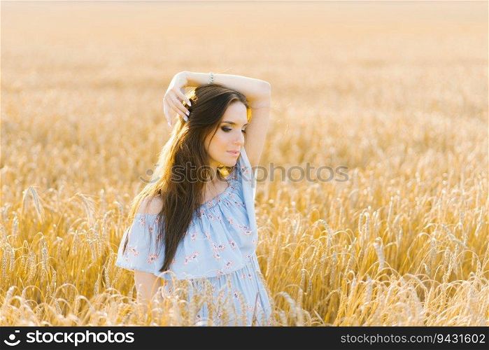 A young woman in a blue dress stands in a field of rye at sunset