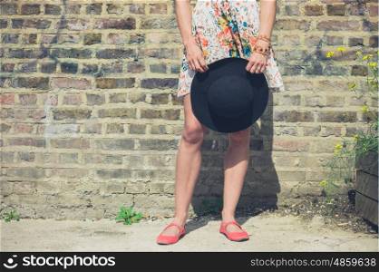 A young woman in a summer dress is standing by a brick wall outside and is holding a hat in her hand