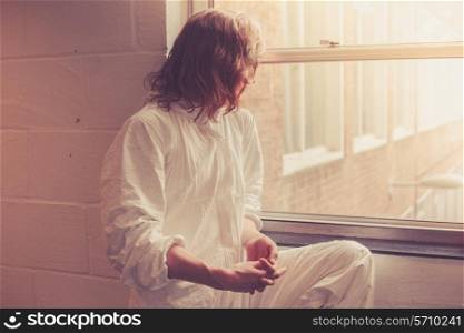 A young woman in a white boiler suit is sitting by a window