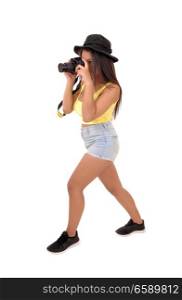 A young woman in shorts and a hat standing in the studio and takingpictures with her camera, isolated over white background.