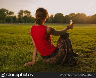 A young woman is drinking from a bottle while sitting on the grass in a park and admiring the sunset