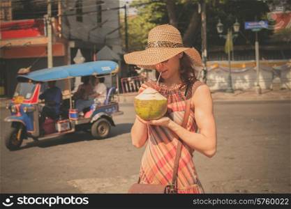 A young woman is drinking from a coconut in the street of an Asian country with a tuktuk in the background