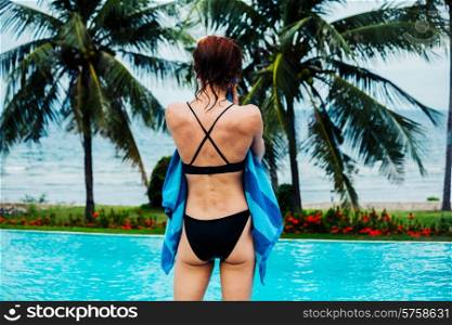 A young woman is drying herself with a towel by a swimming pool with palm trees and the ocean in the background