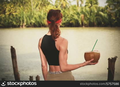 A young woman is holding and drinking from a coconut by a river in the tropics