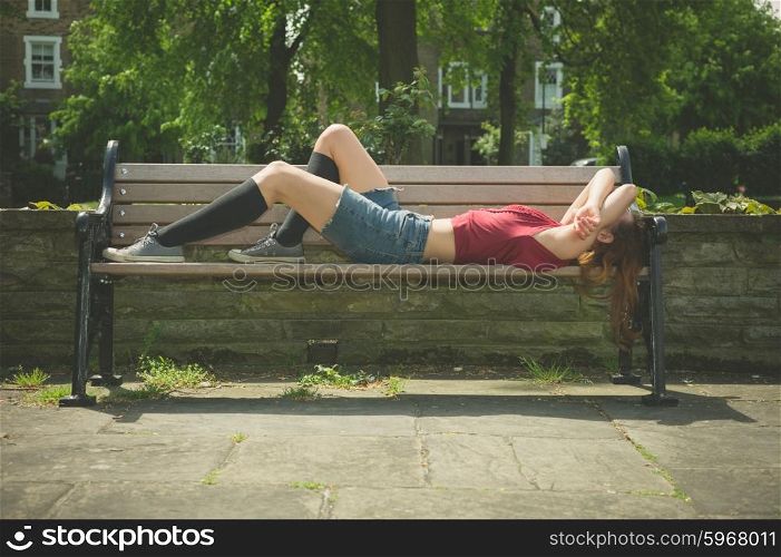 A young woman is lying down and relaxing on a park bench on a sunny summer day