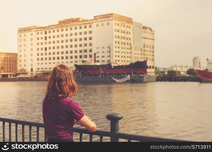 A young woman is relaxing by a river and looking at a dereclict building across it