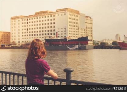 A young woman is relaxing by a river and looking at a dereclict building across it