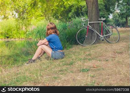 A young woman is relaxing by the water in a park on a summer day with her bicycle resting against a tree in the background
