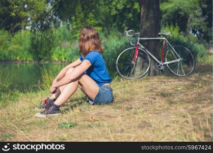 A young woman is relaxing by the water in a park on a summer day with her bicycle resting against a tree in the background