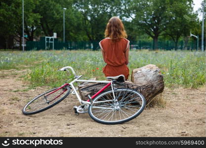 A young woman is sittin gon a log in a meadow with her bike next to her