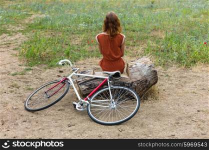 A young woman is sittin gon a log in a meadow with her bike next to her
