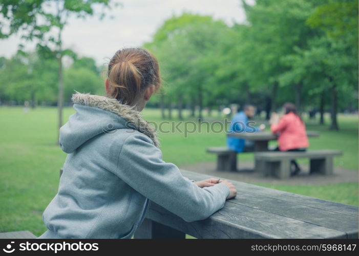 A young woman is sitting alone at a table in the park and is wathing a couple on a picnic