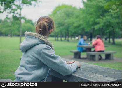 A young woman is sitting alone at a table in the park and is wathing a couple on a picnic