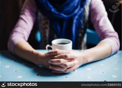 A young woman is sitting at a table with her hands folded around a cup of coffee