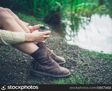 A young woman is sitting by a pond in the forest and is having a cup of tea