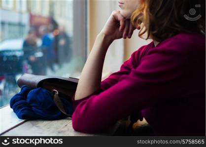 A young woman is sitting by a window and is looking out and day dreaming