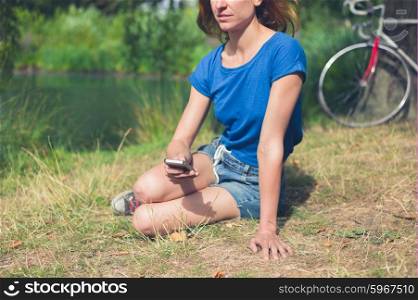 A young woman is sitting by the water in a park and is using a smart phone