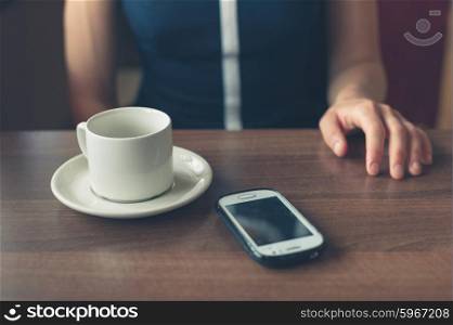 A young woman is sitting by the window in a diner with a cup and a smartphone in front of her