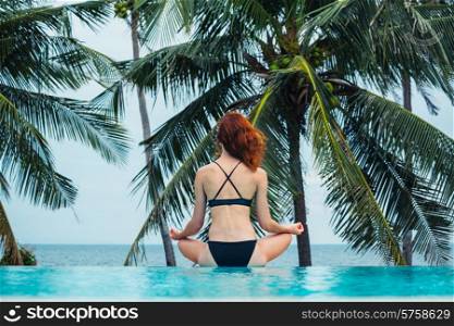 A young woman is sitting in a meditating pose by a swimming pool with palm trees and the ocean in the background