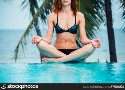 A young woman is sitting in a meditating pose by a swimming pool with palm trees and the ocean in the background