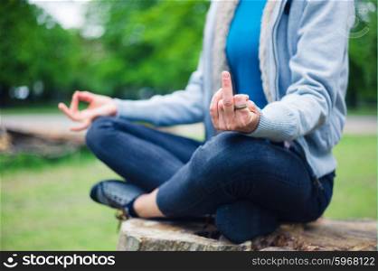 A young woman is sitting in a meditation pose on a tree trunk in the park and is displaying a rude gesture wth her middle finger