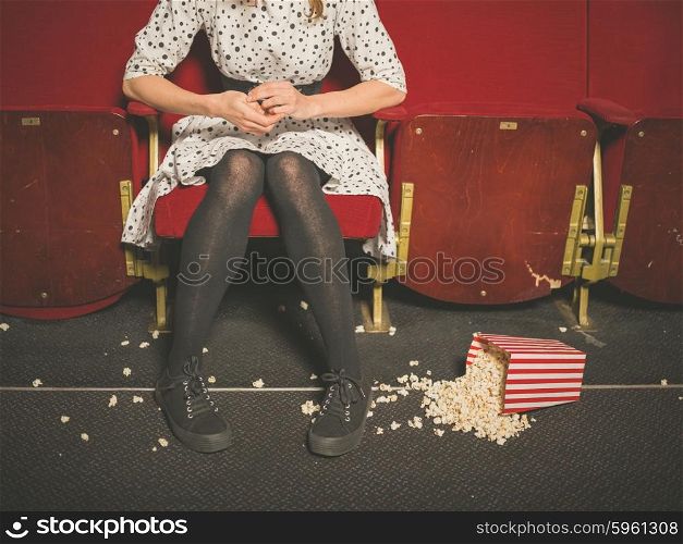 A young woman is sitting in a movie theater with a bucket of popcorn spilled on the floor in front of her