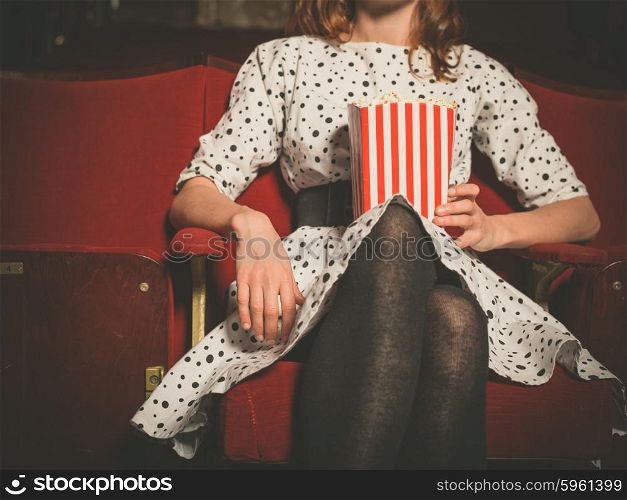 A young woman is sitting in a movie theater with a bucket of popcorn