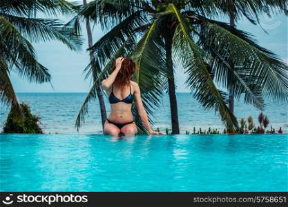 A young woman is sitting in a swimming pool with palm trees and the ocean in the background