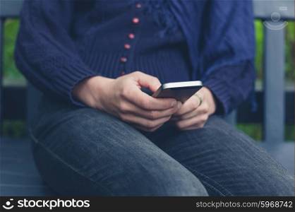 A young woman is sitting on a bench outside in nature and is using a smart phone