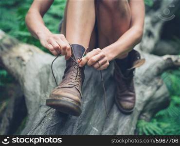 A young woman is sitting on a log in the forest and is tying her boots