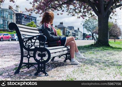 A young woman is sitting on a park bench with cherry blossom on the ground