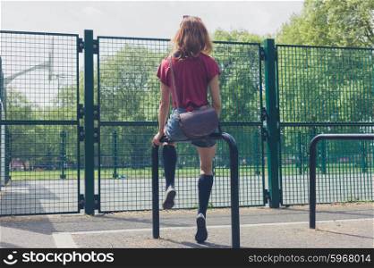 A young woman is sitting on a rail in a park outside a basketball court on a summer day