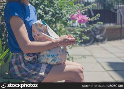 A young woman is sitting on a wall by some flowers and is using her smart phone on a sunny day