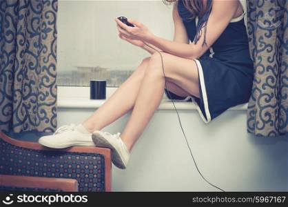 A young woman is sitting on a window sill and is charging her smartphone