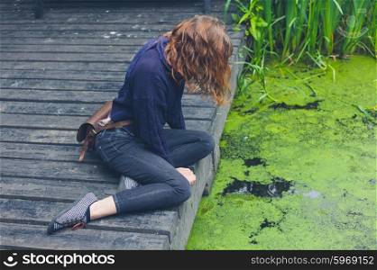A young woman is sitting on a wooden deck by a dirty pond with algae in a forest