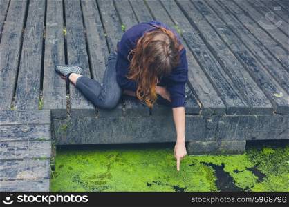 A young woman is sitting on a wooden deck by a pond in a forest and is pointing at something in the water