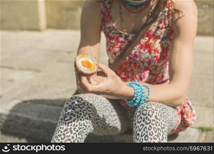 A young woman is sitting on the curb on the street and is eating a scotch egg