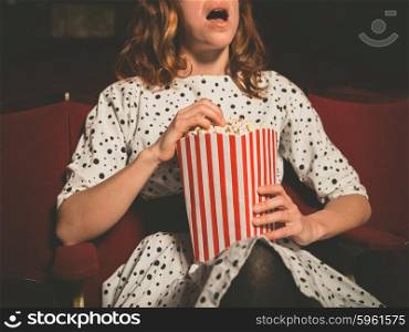 A young woman is sitting on the front row in a movie theater and is watching an exciting film while eating popcorn