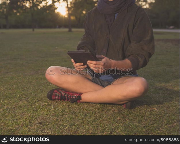 A young woman is sitting on the grass in a park and is using a tablet computer at sunset