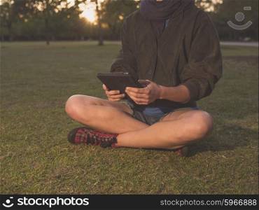 A young woman is sitting on the grass in a park and is using a tablet computer at sunset