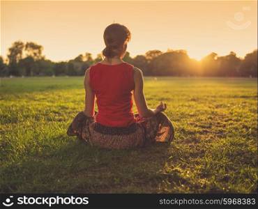 A young woman is sitting on the grass in a park and is meditating at sunset