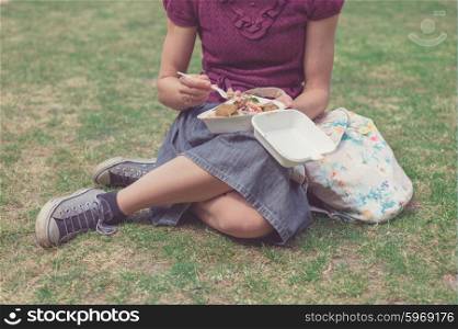 A young woman is sitting on the grass in a park and is eating falafel