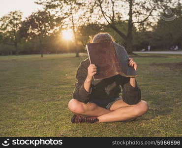 A young woman is sitting on the grass in a park at sunset and is hiding her face behind a notebook