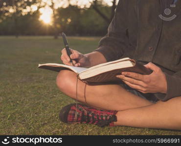 A young woman is sitting on the grass in a park at sunset and is writing in a notebook