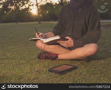 A young woman is sitting on the grass in a park at sunset and is writing in a notebook and using a tablet computer