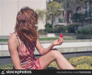 A young woman is sitting on the grass outside an apartment block with a strawberry in her hand